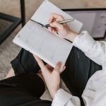 Side view of faceless woman in casual clothes taking notes on notepad while sitting in lotus pose on floor in modern apartment during daytime